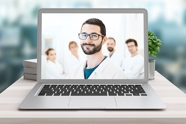 Ein Laptop steht auf einem Tisch. Der Bildschirm zeigt ein Team von Ärzten, wobei ein dunkelhaariger Mann mit Brille im Vordergrund steht und in die Kamera lächelt.